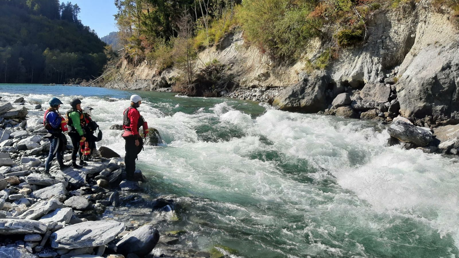 Eine Gruppe Wildwasser-Kanuten steht am Ufer und bespricht, welches an der Wildwasser-Stelle die beste Route ist.